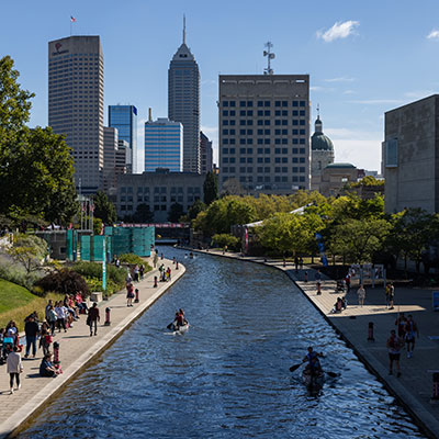 Scene of the Downtown Indianapolis Canal with the city skyline in the background, two boats on the water and spectators on either side of the canal.