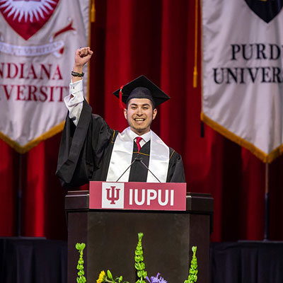 IUPUI Commencement student speaker Jonathan Zachary Madrigal in cap and gown at podium during commencement with fist raised as if in victory.  Behind him part of the Indiana University and Purdue University banners are visible.  