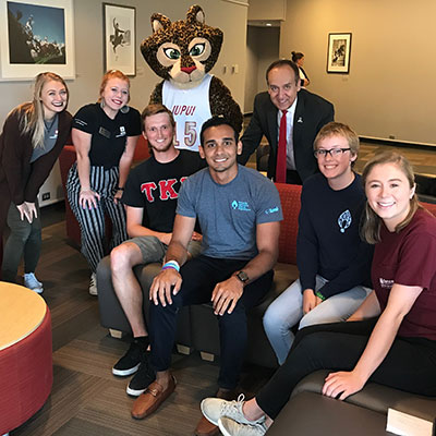 Students wearing IUPUI gear are seated in front of Chancellor Paydar and IUPUI mascot Jazzy.  All are smiling for the camera. 