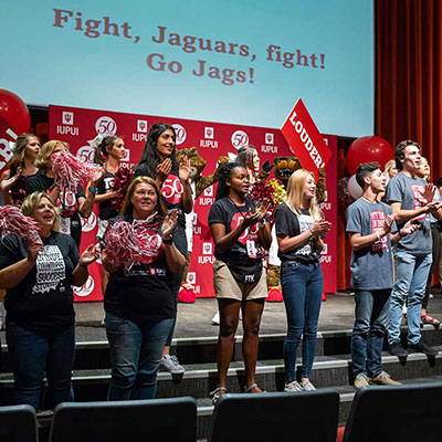 IUPUI students wearing 50th Anniversary t-shirts, holding pom pons and signs stand on a stage with an IUPUI backdrop in the background and on a screen above their heads the words "Fight, Jaguars, fight! Go Jags!" are visible.
