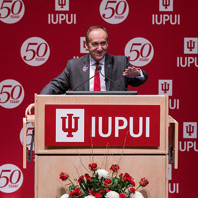 Chancellor Nasser Paydar stands at IUPUI-branded podium reaching out to audience.  In the background an IUPUI 50th anniversary backdrop is visible, and red and white flowers appear in front of the podium. 