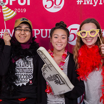 Three female IUPUI students appear in front of an IUPUI 50th Anniversary background with celebratory props, including a giant foam finger with #1 on it.  