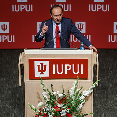 Chancellor Nasser Paydar at IUPUI-branded podium with IUPUI background in back of him and red and white flowers in front of the podium. 