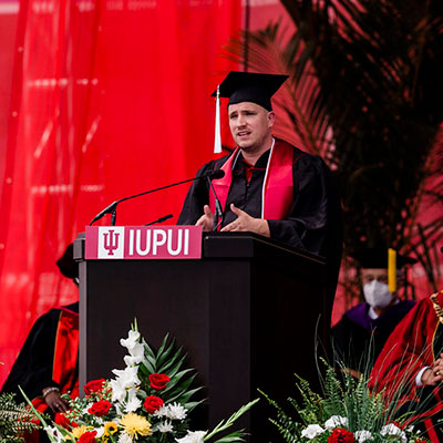 IUPUI undergraduate student speaker Matt Brown at commencement ceremony.  He is wearing academic regalia, standing behind the IUPUI podium.  Behind him is a red theater curtain and in front of the podium is a spray of white, red, and yellow flowers.