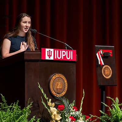 Student representative Emily Wolf speaks at Ball Hall Rededication.  She is standing behind an IUPUI-branded podium, which also features the Indiana University seal.  Behind her is a red theater curtain, and in front of the podium are flowers and greenery.