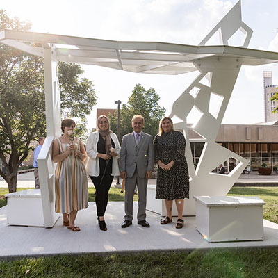 Chancellor Nasser Paydar stands with three Herron School of Art and Design student artists who designed the Skylight, a sculpture, under which the four of them stand.  The day is sunny and trees are visible in the background.